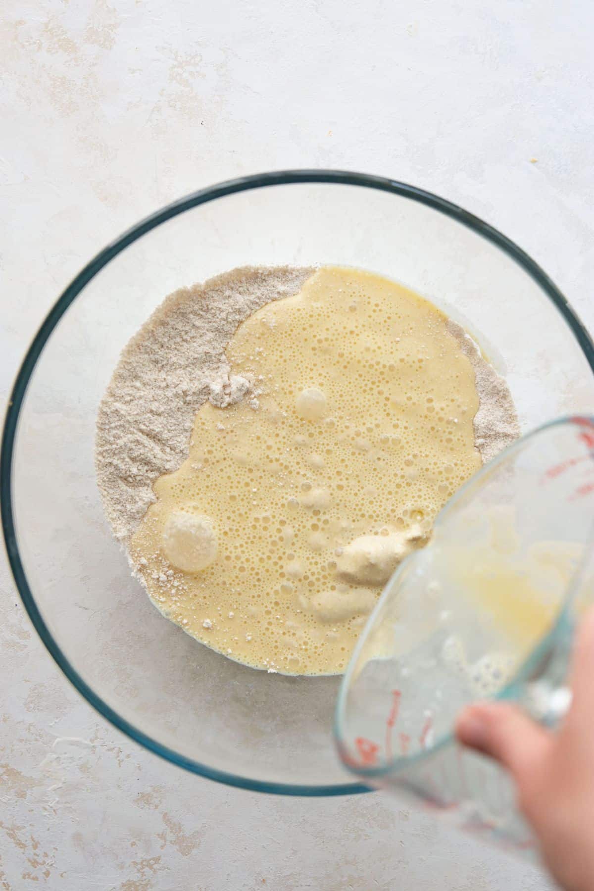 Wet ingredients being poured from a glass measuring cup into dry oat flour mixture in a large glass bowl.