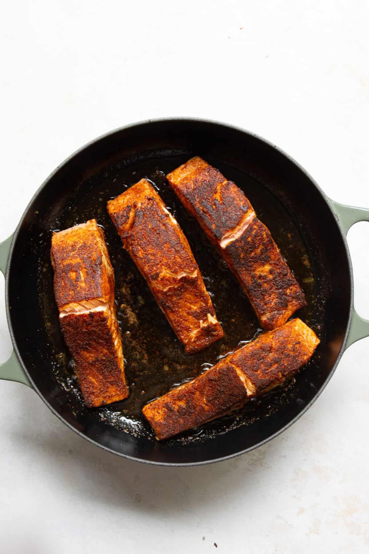 Four blackened salmon fillets seared flesh-side up in a cast iron skillet with a dark, crispy Cajun spice crust.