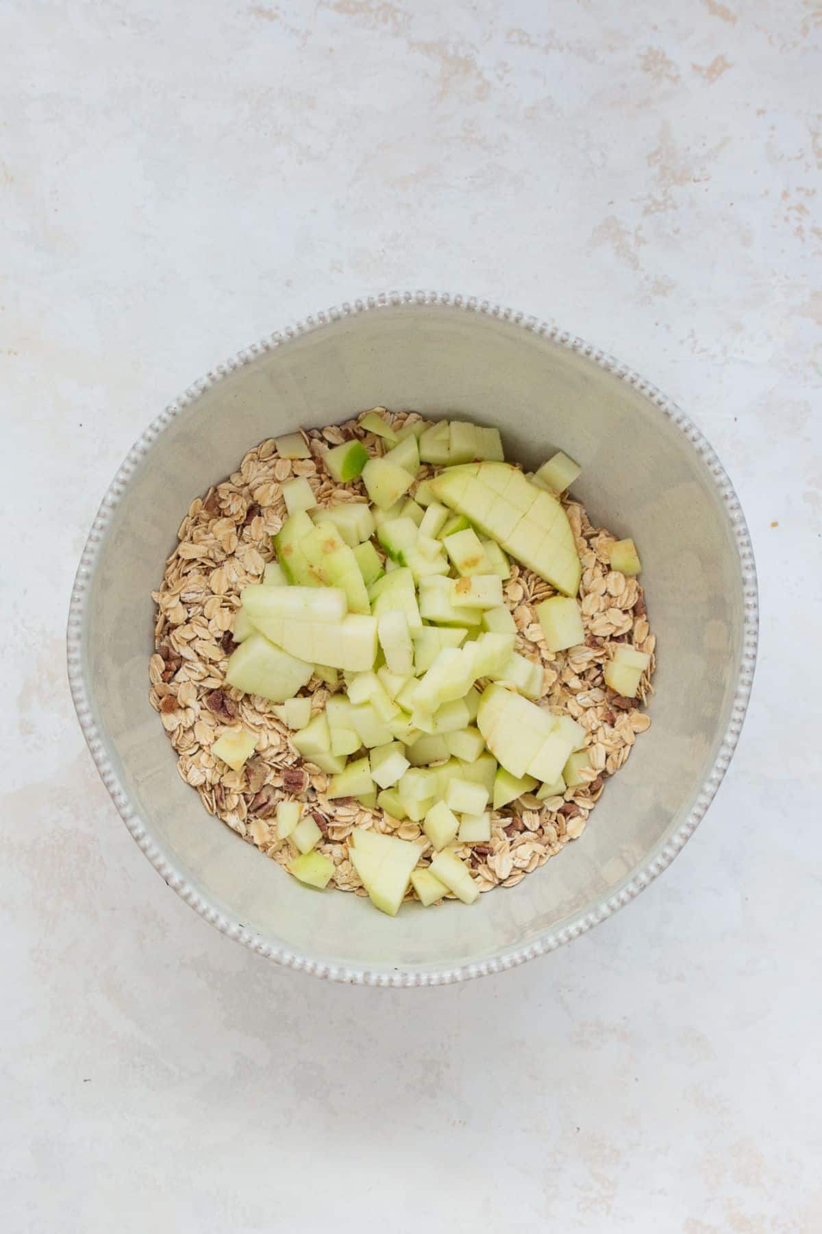 Chopped Granny Smith apples on top of rolled oats and spices in a mixing bowl.
