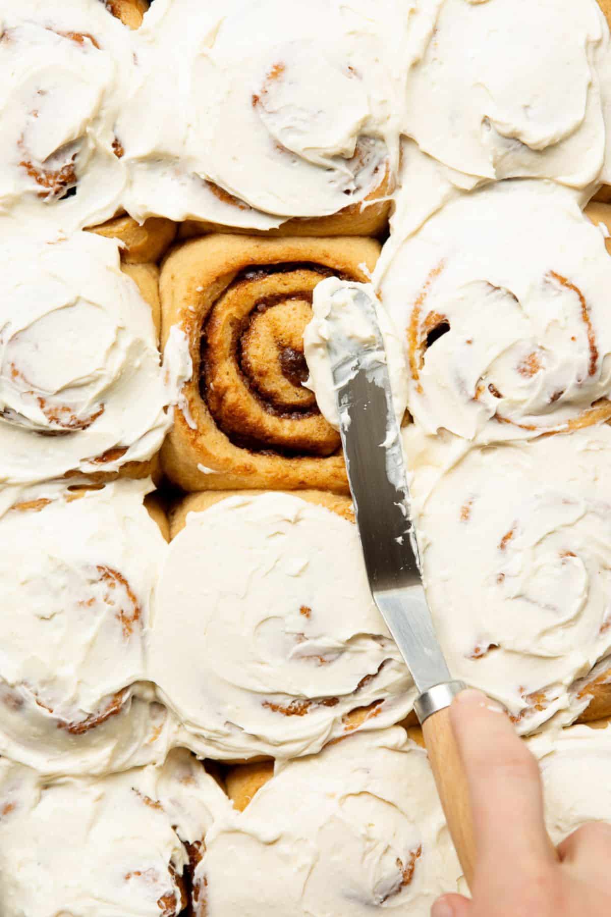 Greek yogurt icing being spread over warm healthy cinnamon rolls in the baking pan with an offset spatula.