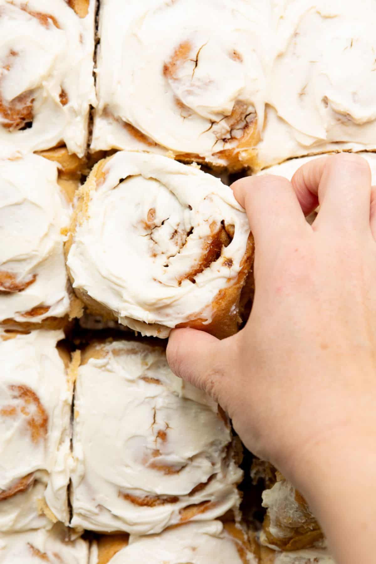 Hand lifting a freshly frosted healthy cinnamon roll out of the pan, with Greek yogurt icing dripping down the sides