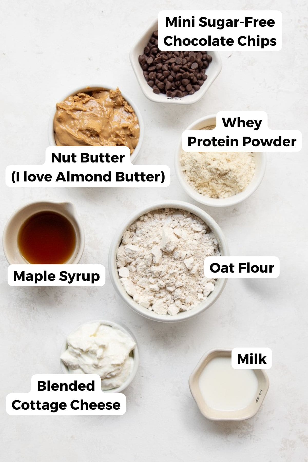 Ingredients for high-protein edible cookie dough including blended cottage cheese, oat flour, whey protein powder, almond butter, maple syrup, milk, and mini sugar-free chocolate chips in small bowls.