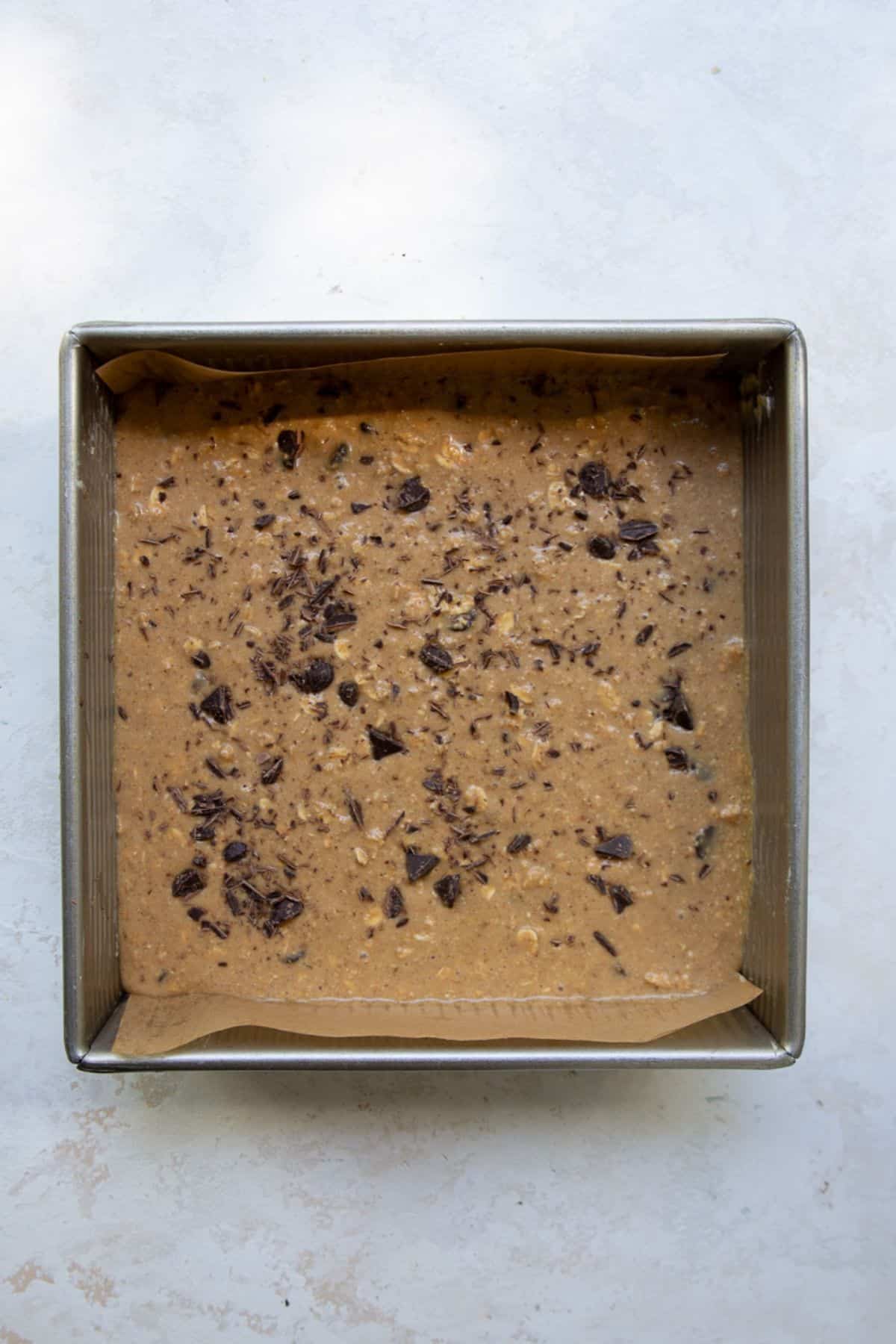 Unbaked healthy chocolate chip oatmeal bar batter spread in a parchment-lined square baking pan, topped with chopped dark chocolate.