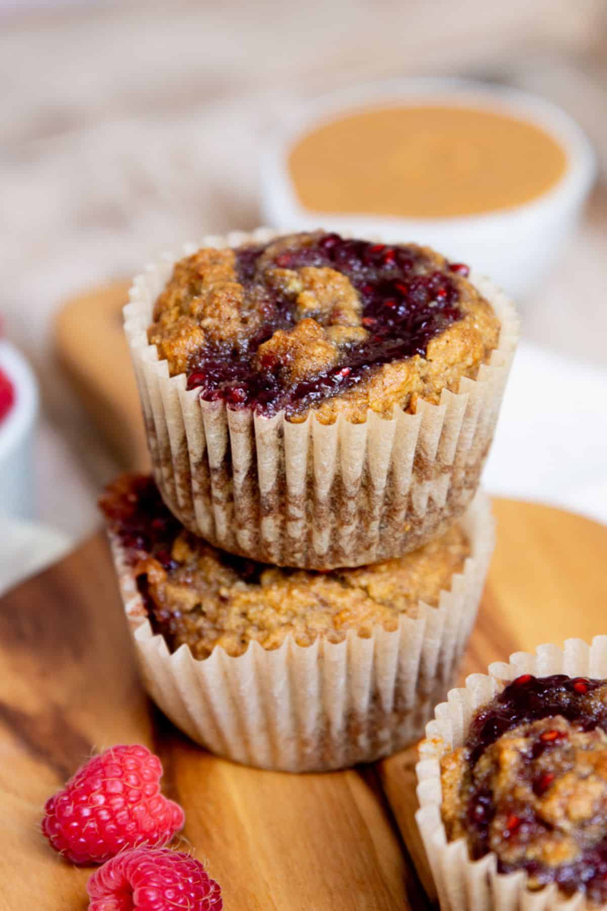 Two gluten-free peanut butter and jelly banana muffins stacked on a wood board with a raspberry jam swirl on top.