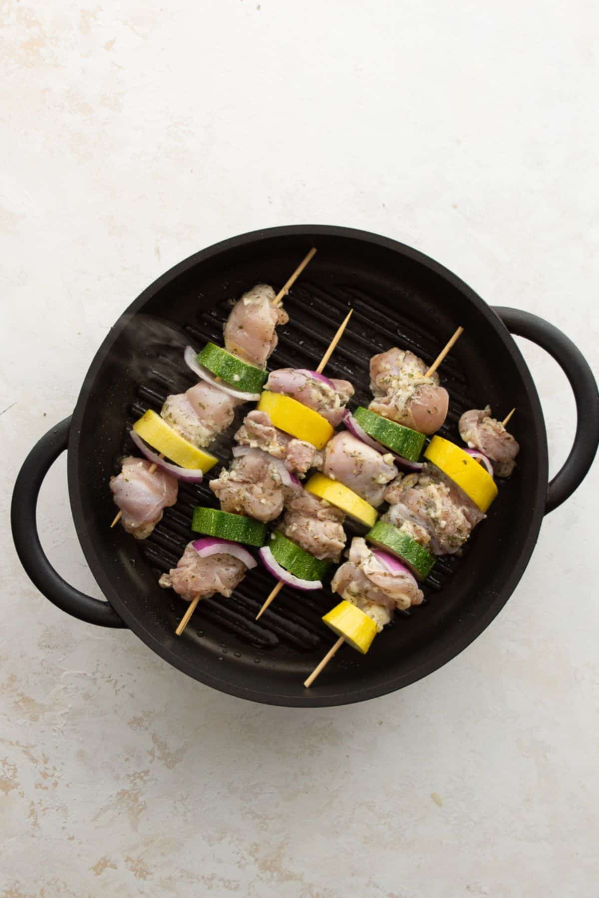 Raw Greek chicken skewers with zucchini and red onion on a preheated grill pan.