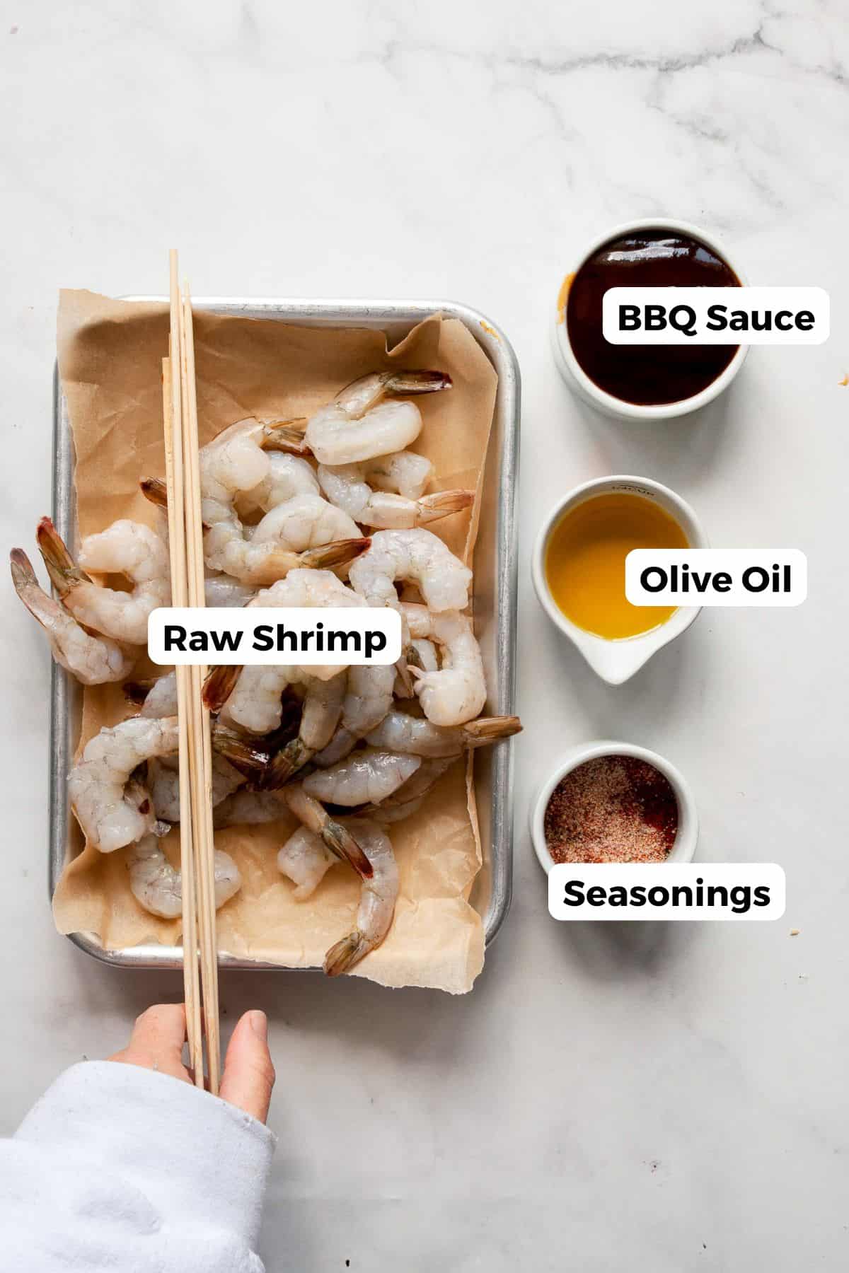All ingredients in BBQ shrimp in bowls labeled, including: raw shrimp, BBQ sauce, olive oil, salt, wooden skewers, and paprika.