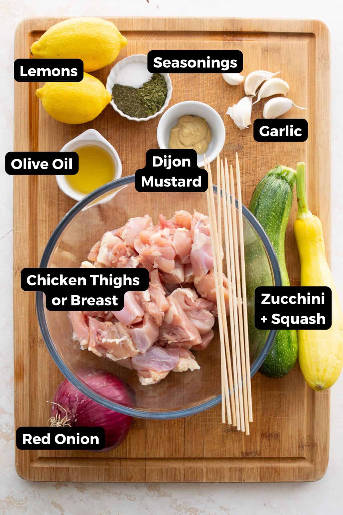 Labeled ingredients for grilled Greek chicken kabobs: chicken thighs, lemons, garlic, olive oil, Dijon, seasonings, zucchini, squash, and red onion.