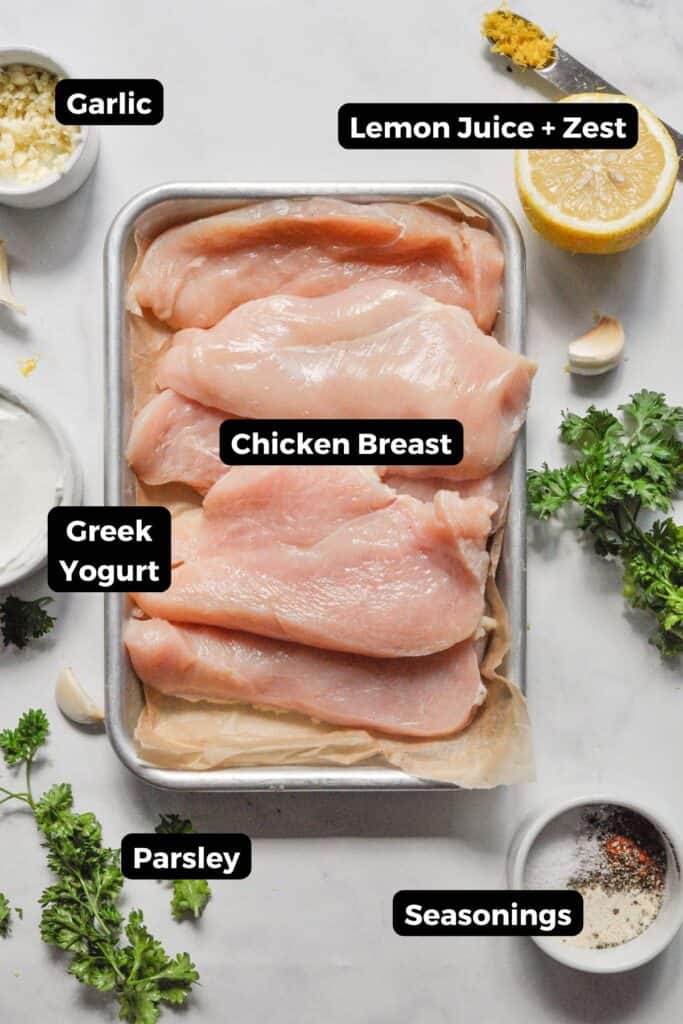 Ingredients for Greek yogurt marinated chicken including raw chicken breasts, lemon, garlic, parsley, and spices.