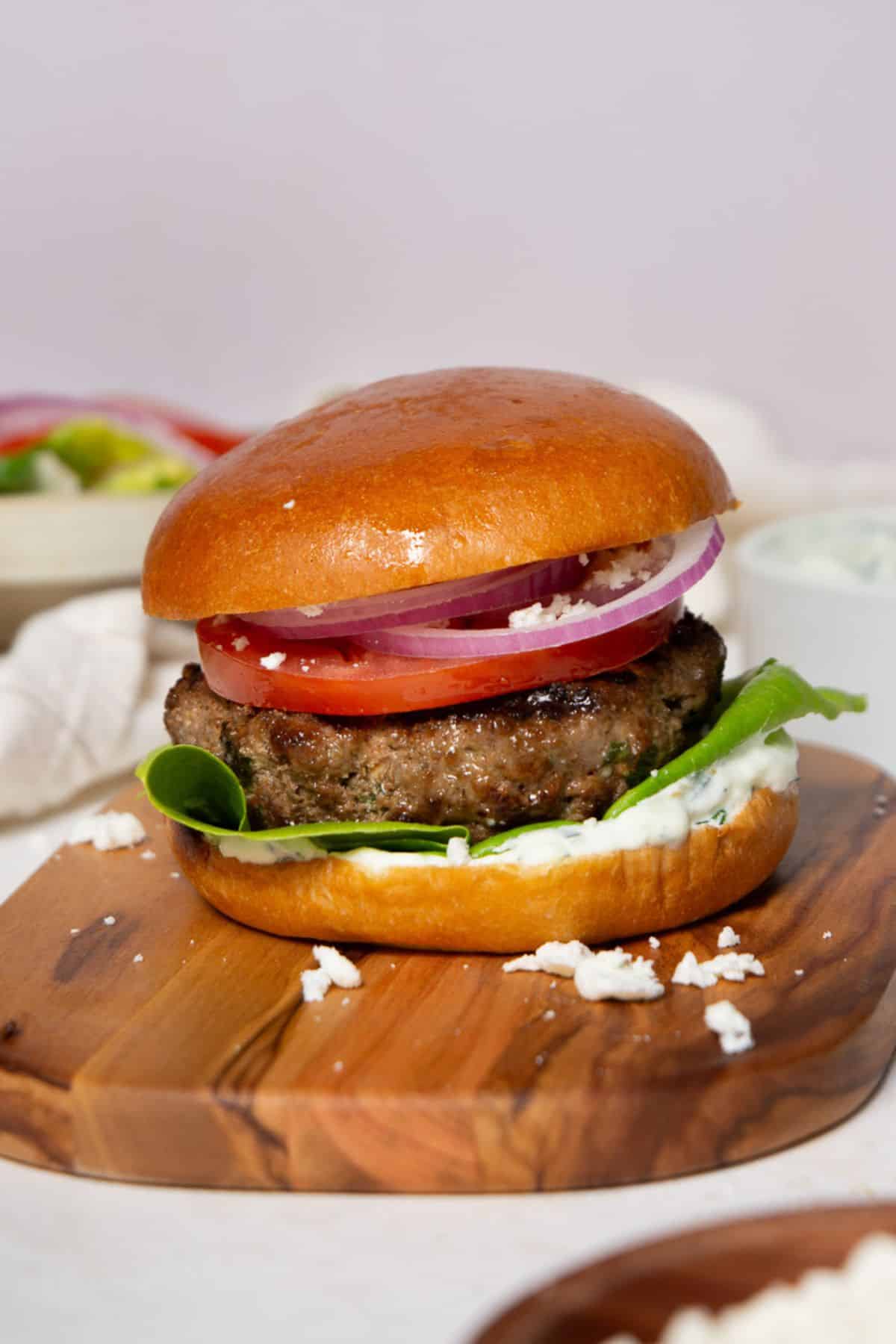 Juicy bison burger on a toasted brioche bun with lettuce, tomato, red onion, tzatziki, and feta on a wooden board.
