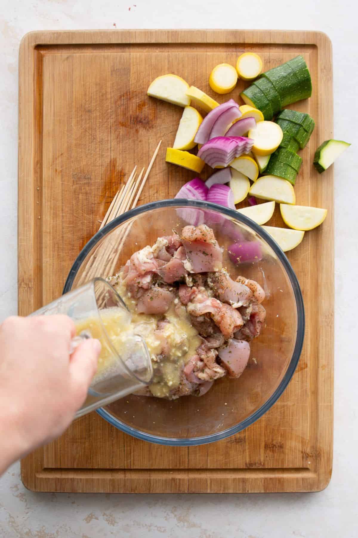Lemon, garlic, and dill marinade being poured over cubed chicken thighs in a glass bowl.