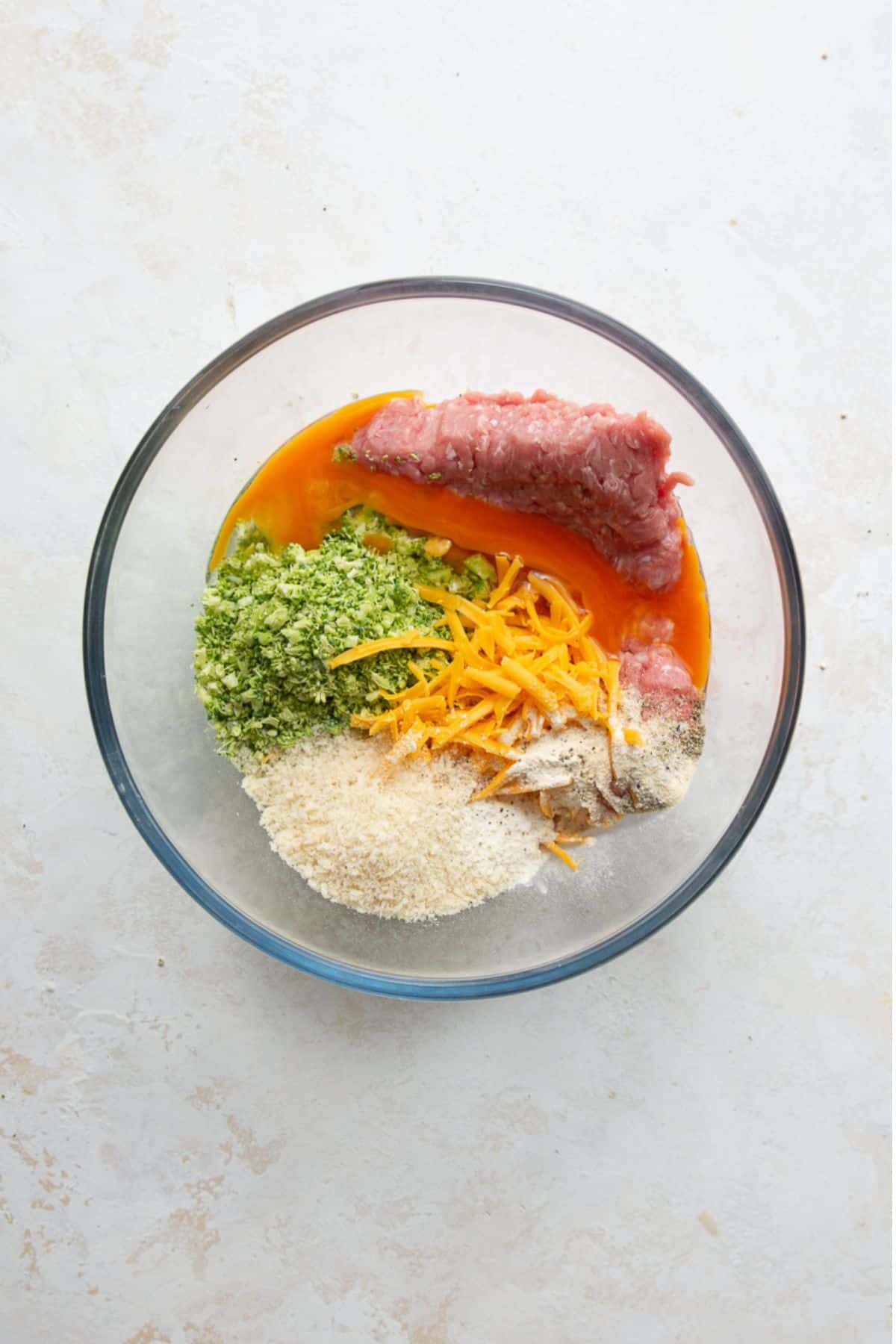 Ground turkey, riced broccoli, shredded cheddar, egg, and panko in a glass bowl before mixing into meatballs.