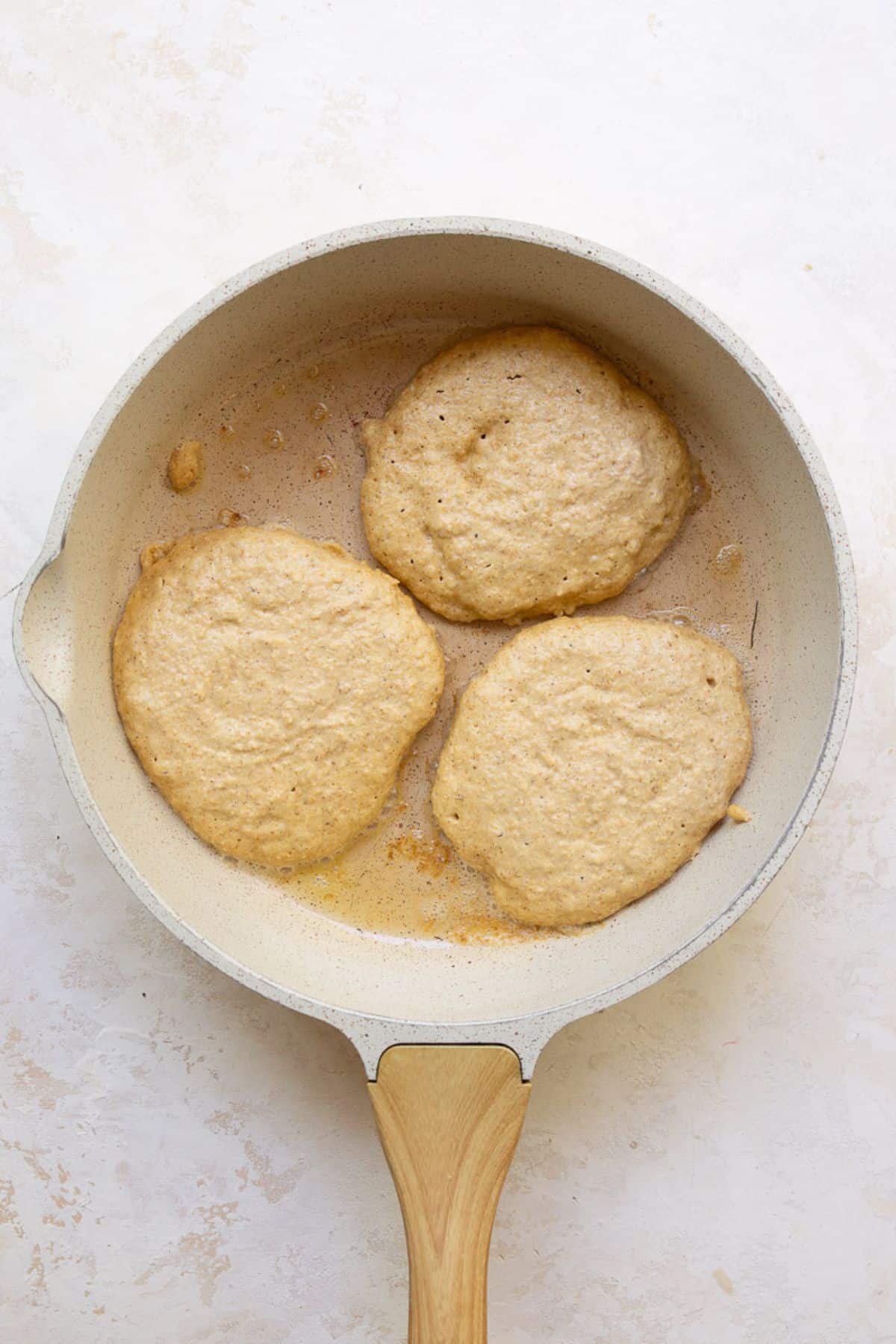 Three protein oatmeal pancakes cooking in a buttered pan before flipping.
