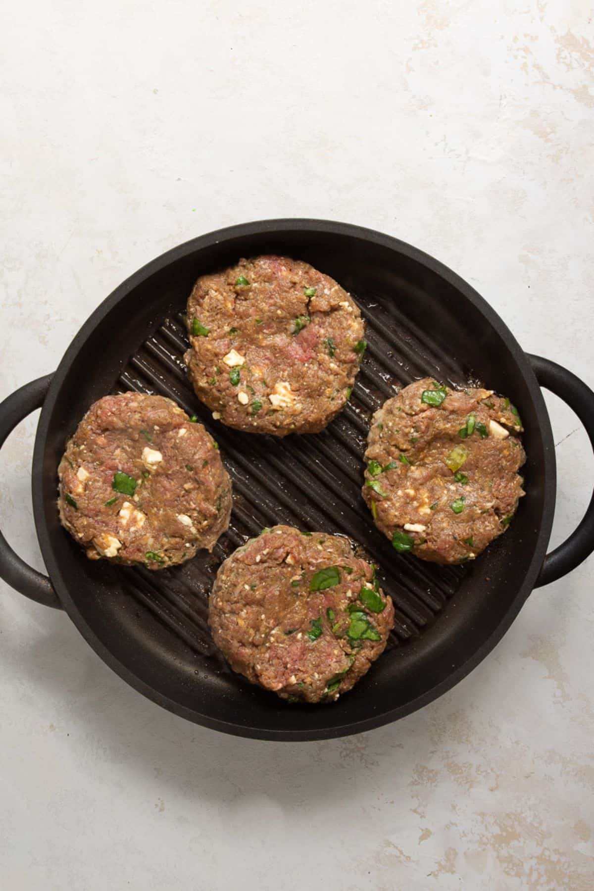 Four raw bison burger patties cooking on a cast iron grill pan.