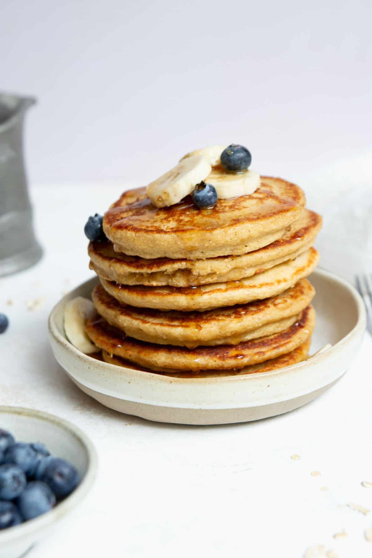 Tall stack of protein oatmeal blender pancakes with banana, blueberries, and maple syrup.