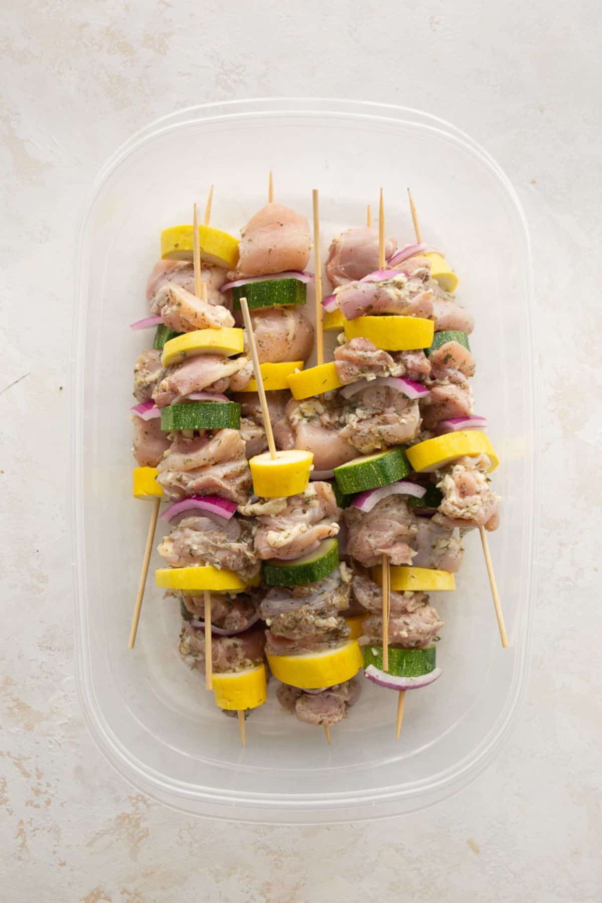 Assembled raw Greek chicken kabobs threaded with zucchini, yellow squash, and red onion in a container.