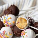 High-protein cake pops on a white plate, dipped in white and dark chocolate with rainbow sprinkles, one bitten to show the soft vanilla center