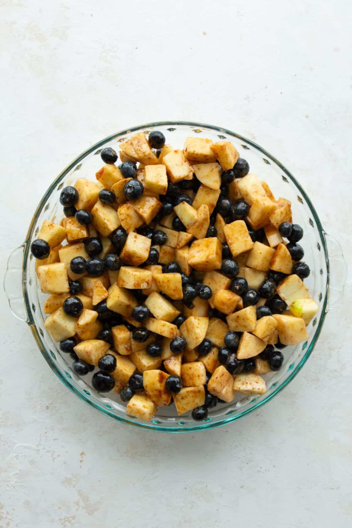Cinnamon-coated apples and blueberries spread in a glass pie dish before baking.