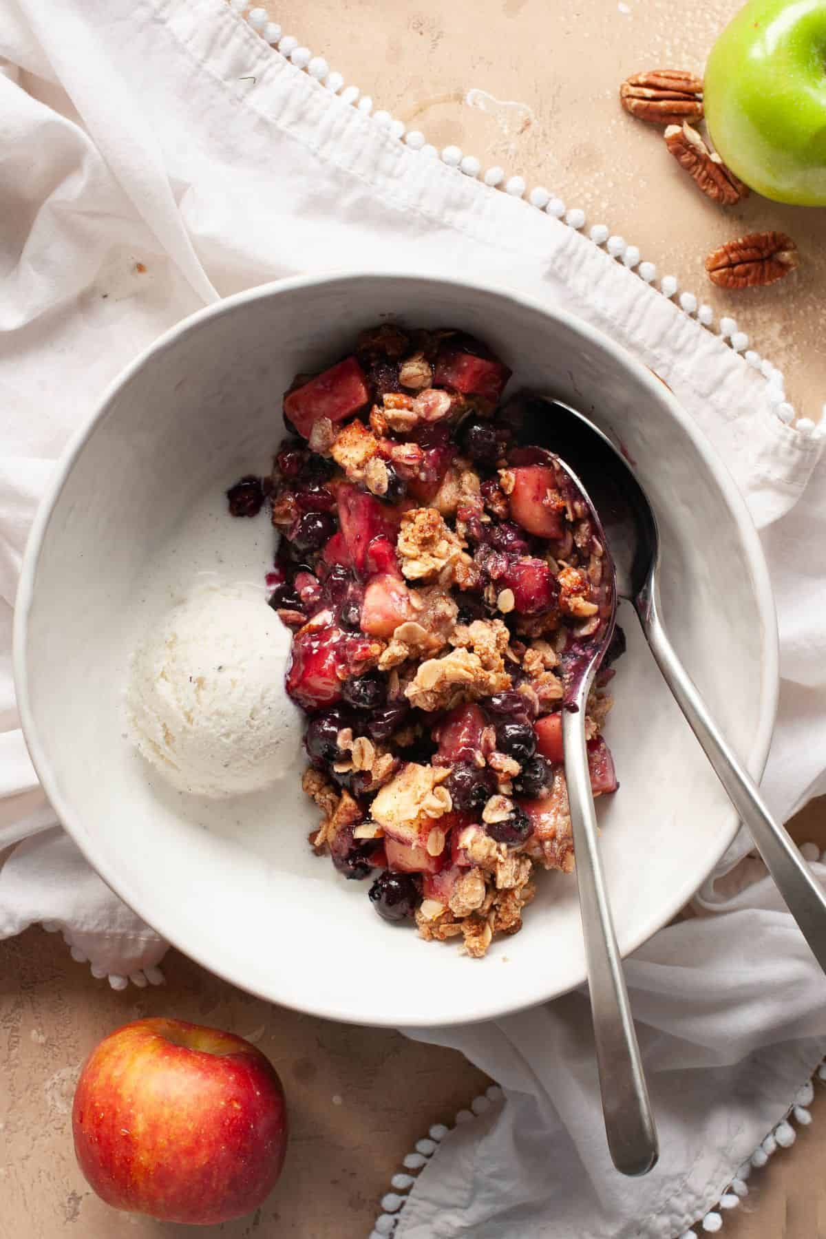 Serving of apple blueberry crisp with vanilla ice cream in a white bowl with a spoon.