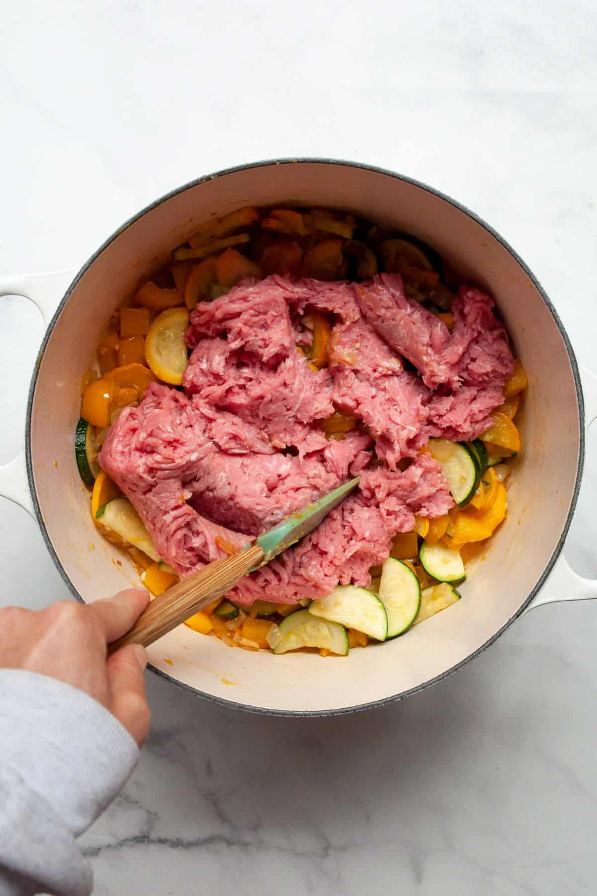 Lean ground turkey added to sautéed vegetables in a Dutch oven, being broken up with a wooden spoon.