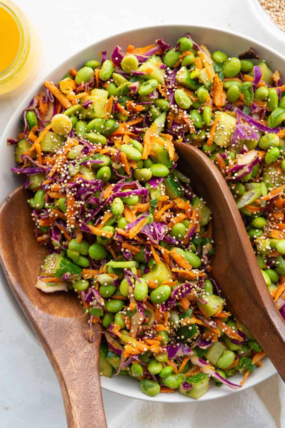 Asian edamame crunch salad topped with puffed quinoa and served with two wooden spoons in a white bowl.