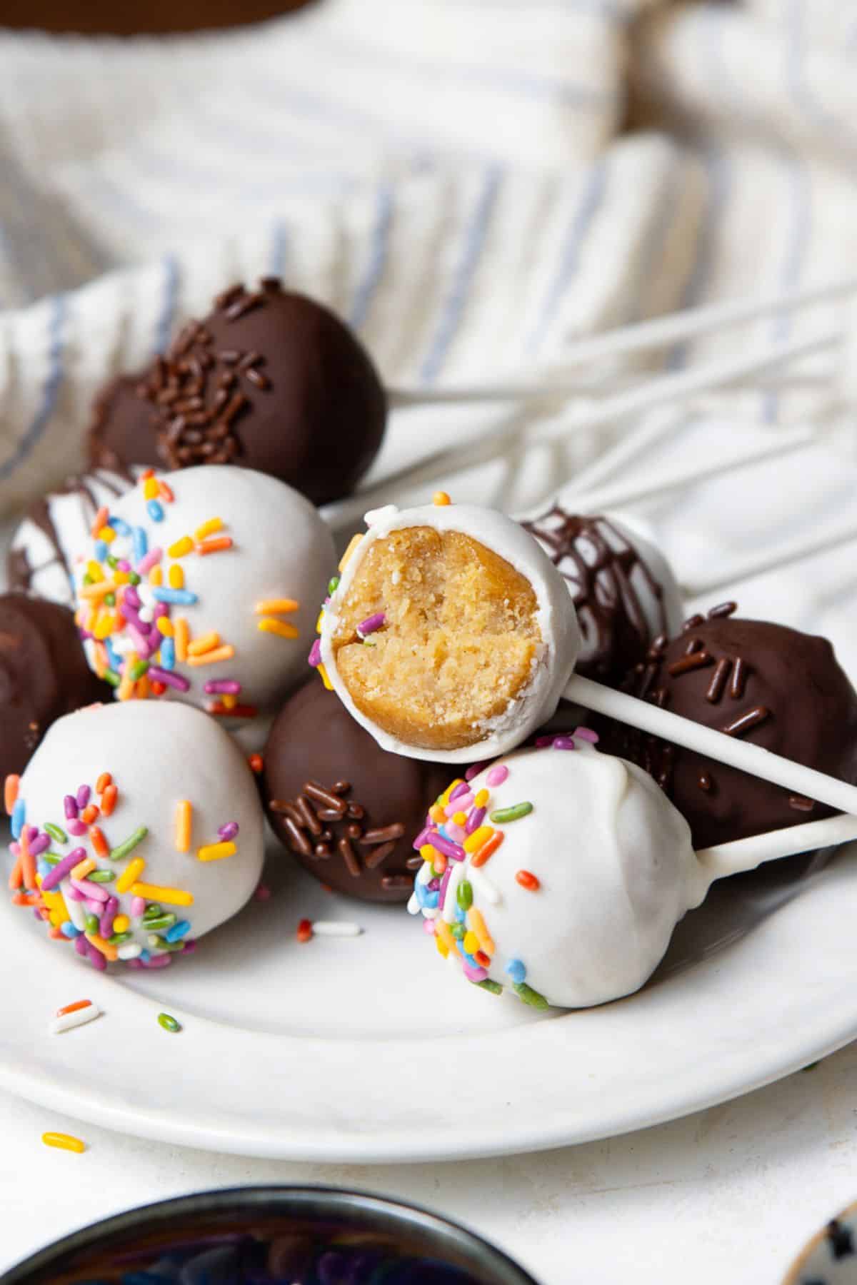 High-protein cake pops made with protein powder on a white plate, dipped in white and dark chocolate with a bitten pop showing the vanilla center.