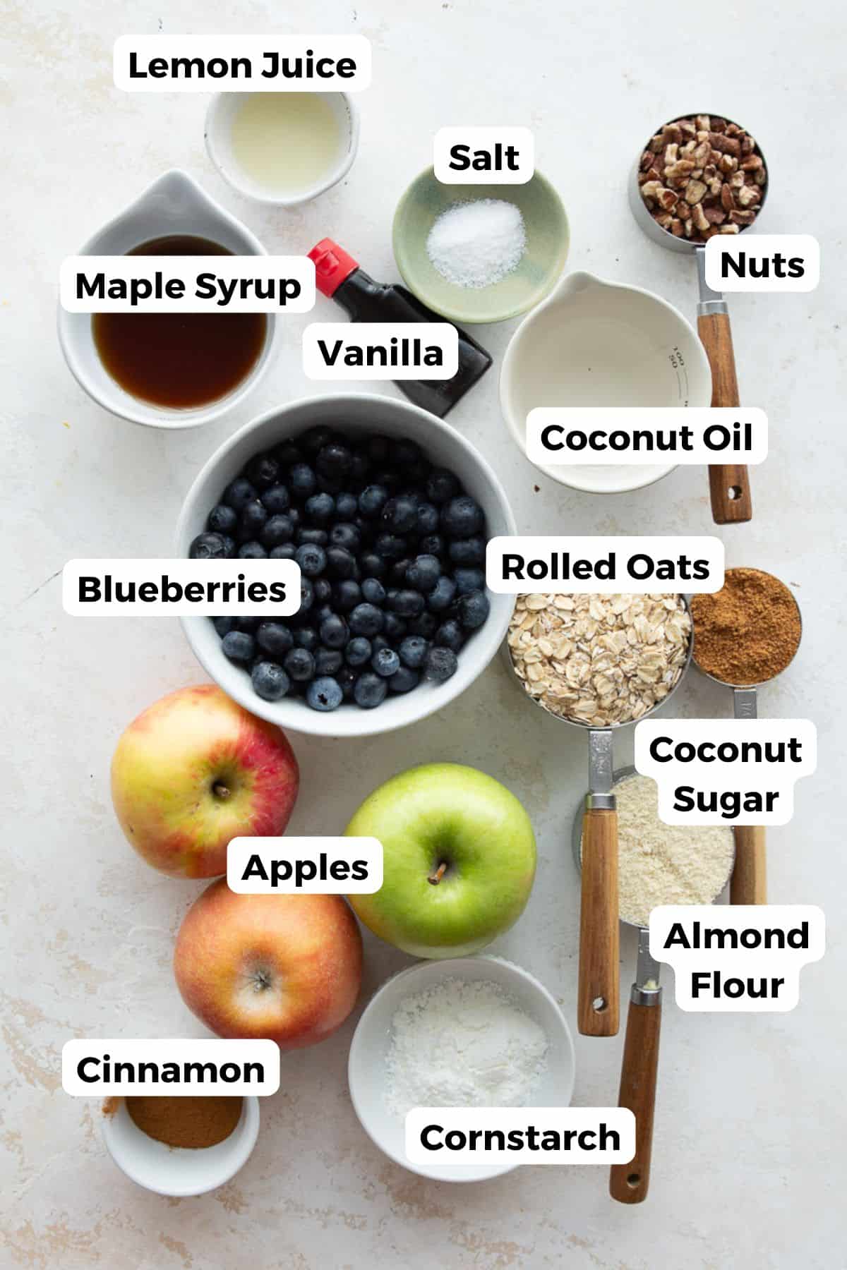 Labeled ingredients for healthy apple blueberry crisp including apples, blueberries, oats, almond flour, and maple syrup.