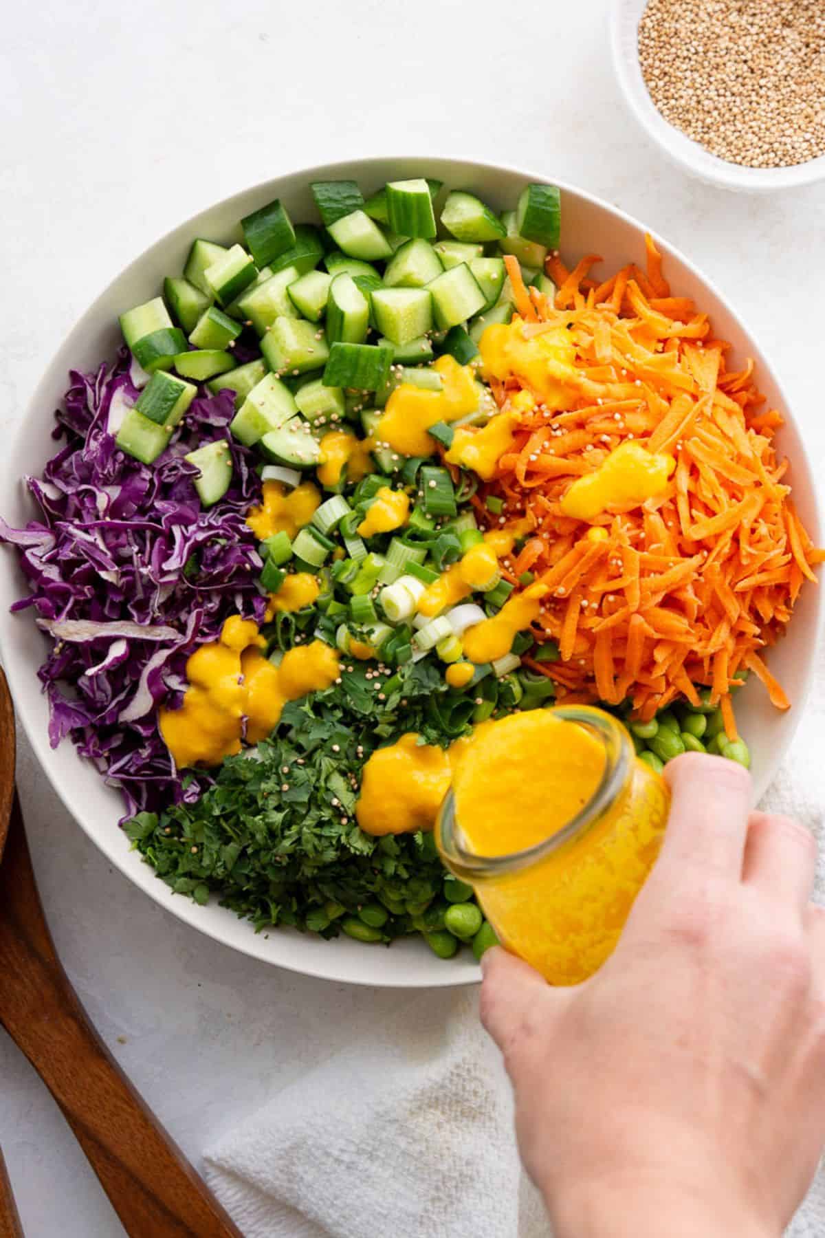 Carrot ginger dressing being poured from a jar over an Asian edamame crunch salad with red cabbage, cucumber, carrots, and cilantro in a white bowl.