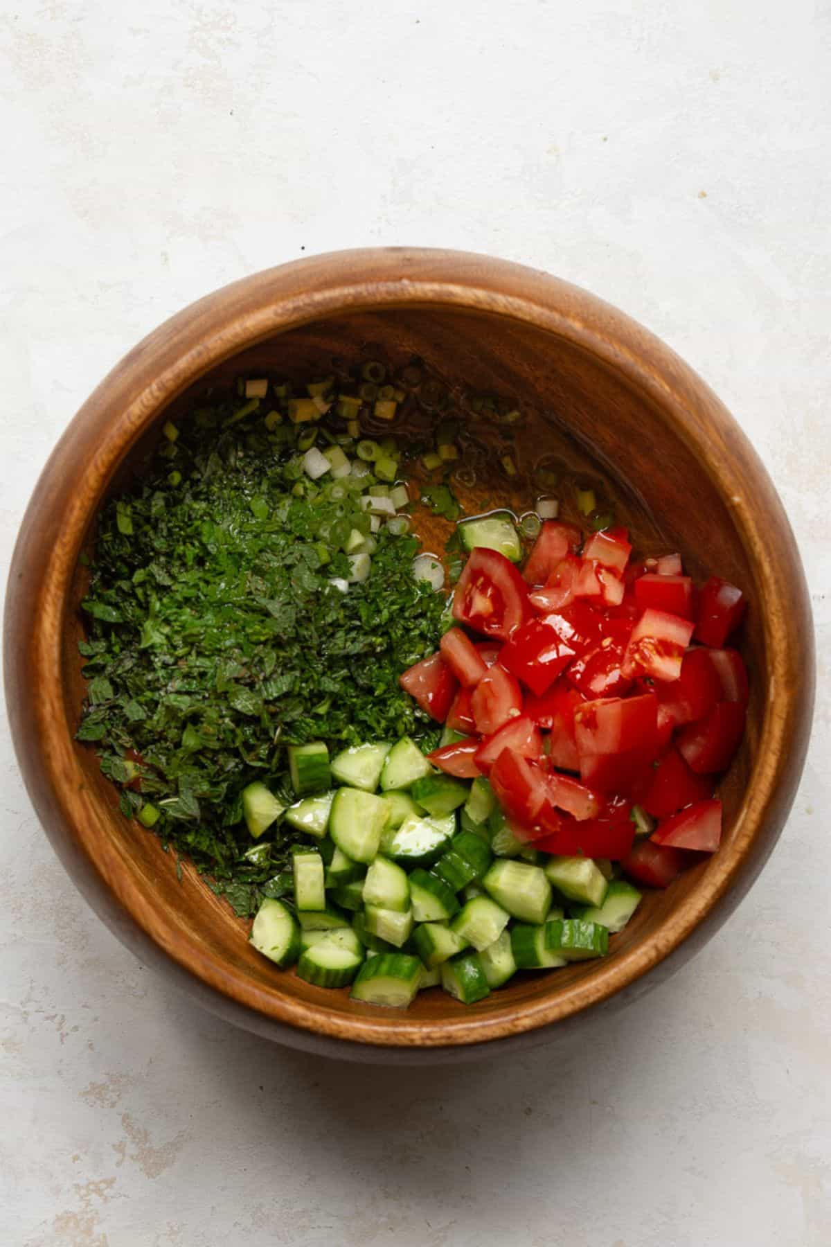 Chopped parsley, mint, scallions, cucumber, and tomato arranged in a wooden bowl.