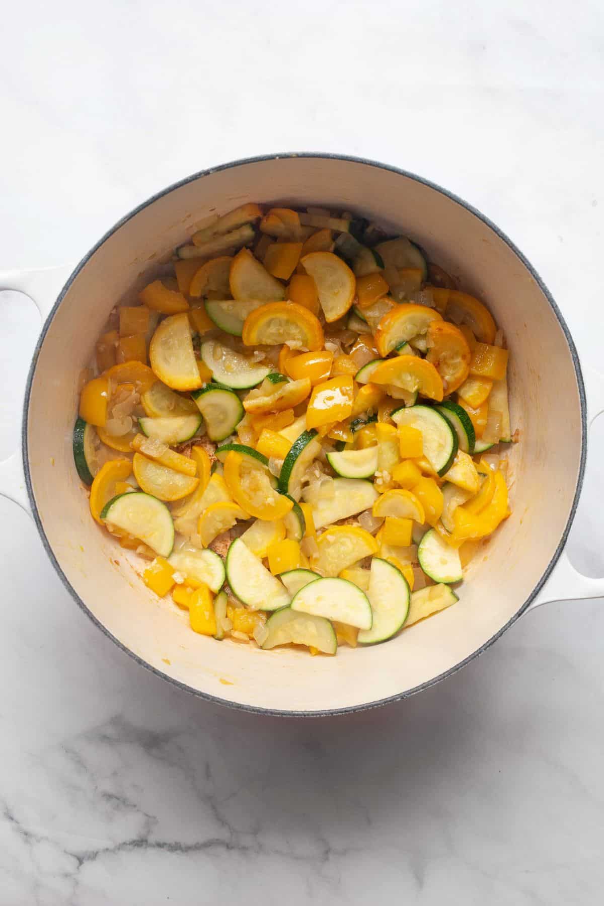 Zucchini, yellow squash, onion, and bell pepper sautéing in a white Dutch oven.