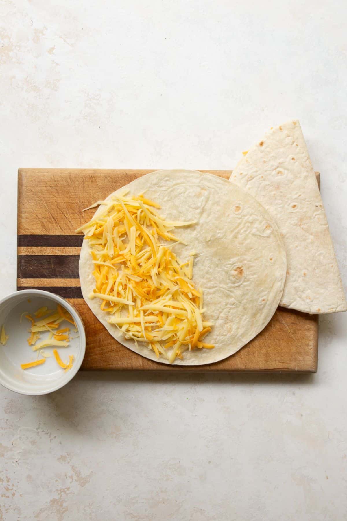 Shredded cheddar spread over half a flour tortilla on a cutting board before folding.