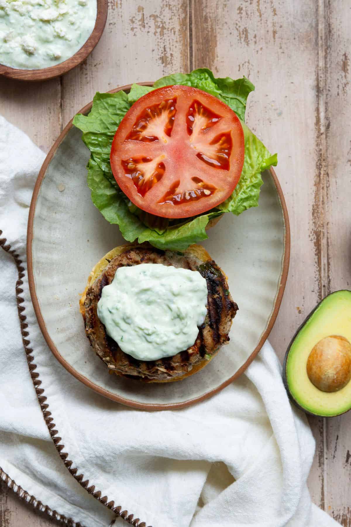 Greek turkey burger on a toasted bun topped with tzatziki, sliced tomato, and lettuce, with avocado alongside.