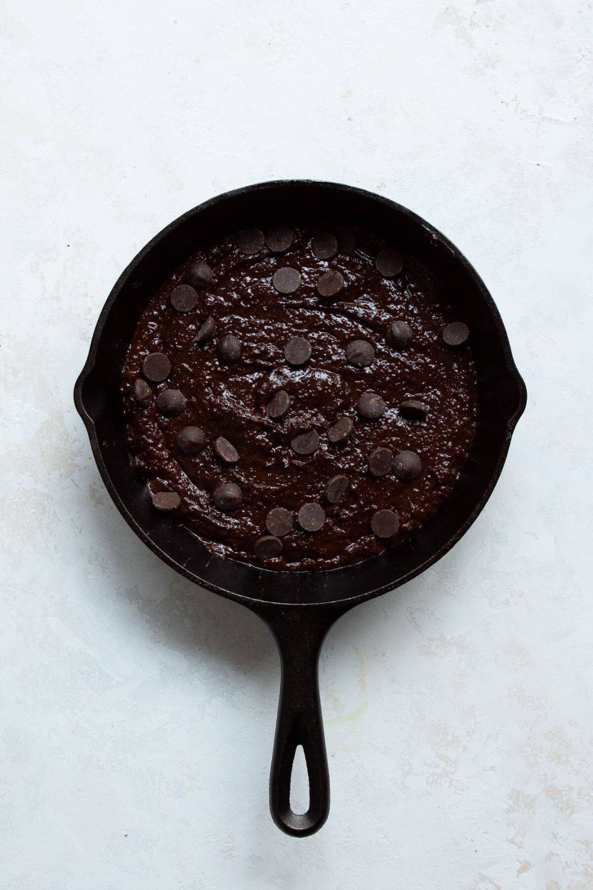 Brownie batter spread into a greased cast iron skillet and topped with chocolate chips.
