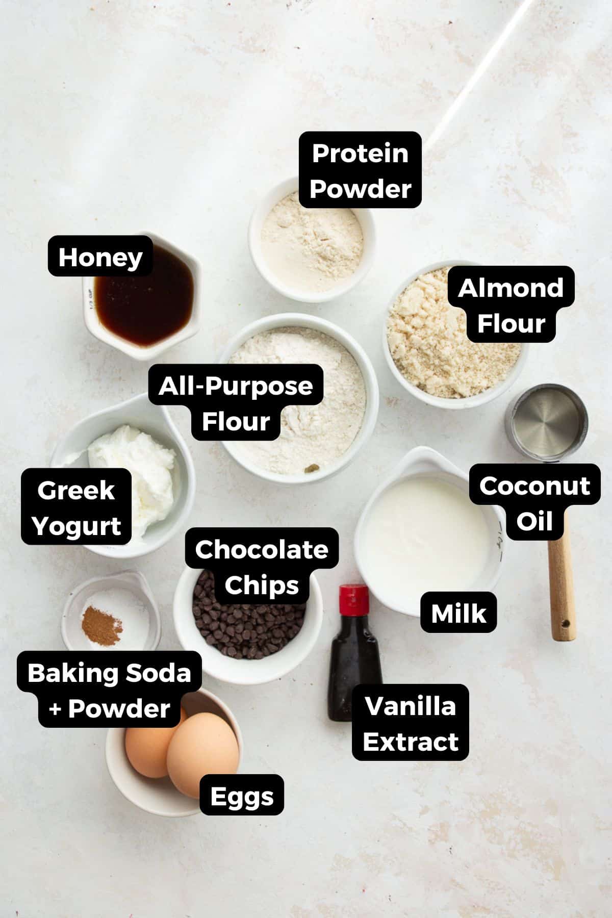 Labeled ingredients for chocolate chip protein muffins including protein powder, almond flour, all-purpose flour, Greek yogurt, honey, coconut oil, milk, eggs, and mini chocolate chips.