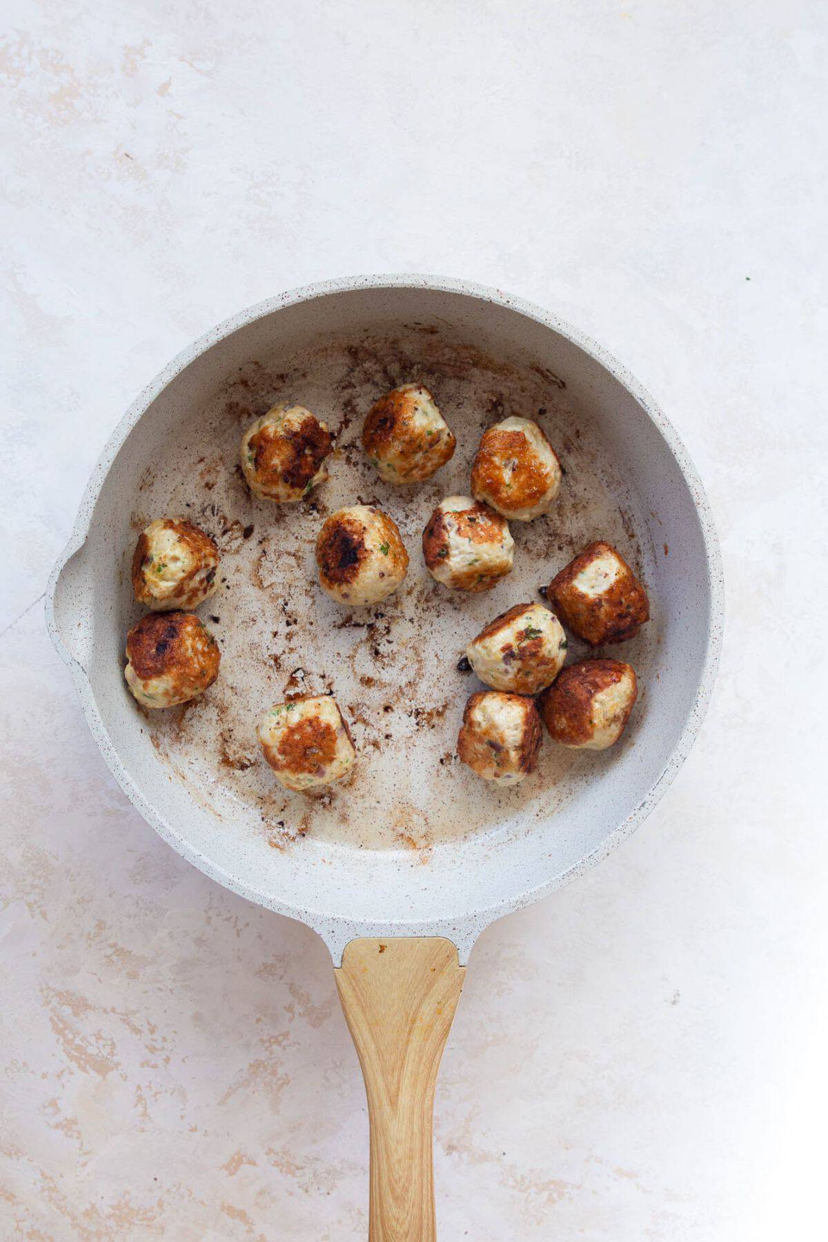 Greek chicken meatballs browning in a white skillet, golden on the seared sides.