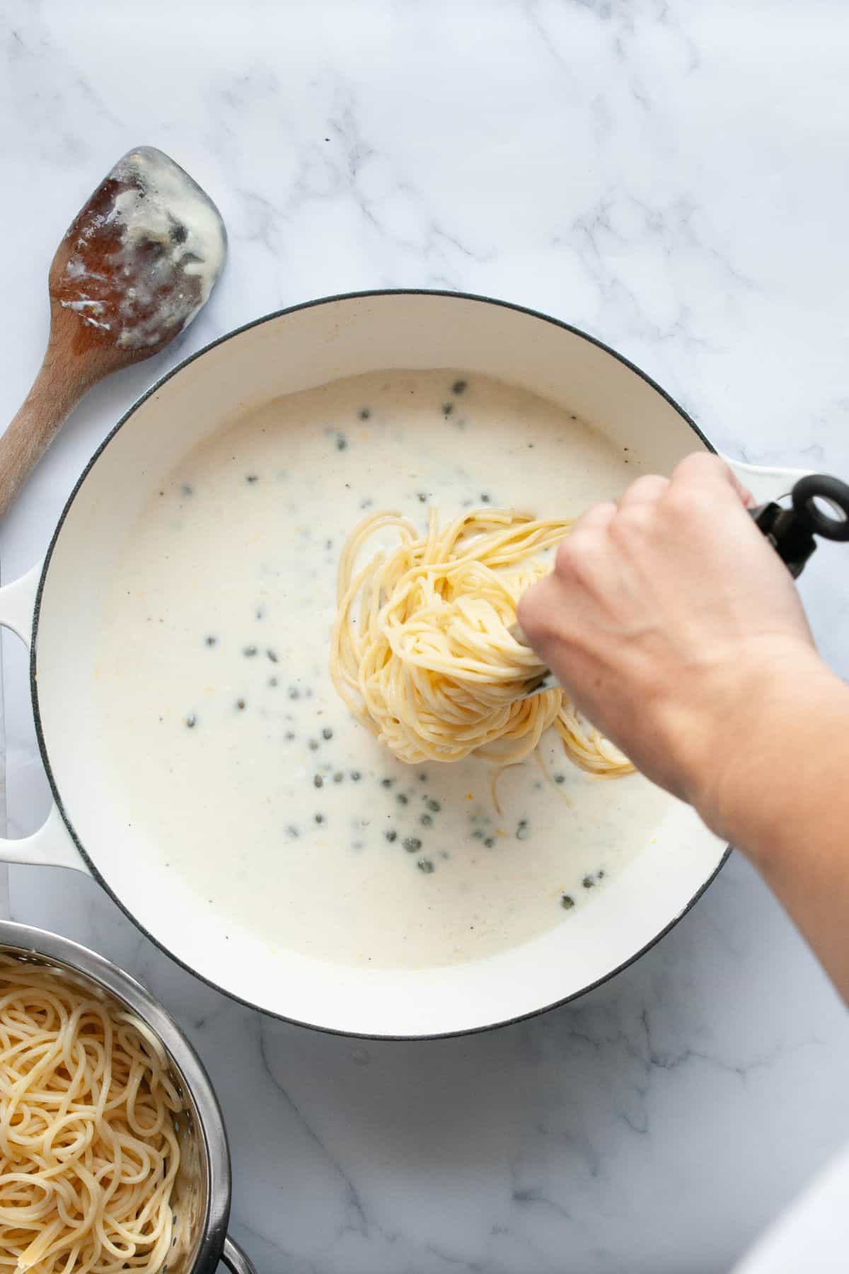 Tongs lowering spaghetti into creamy lemon caper sauce with capers.