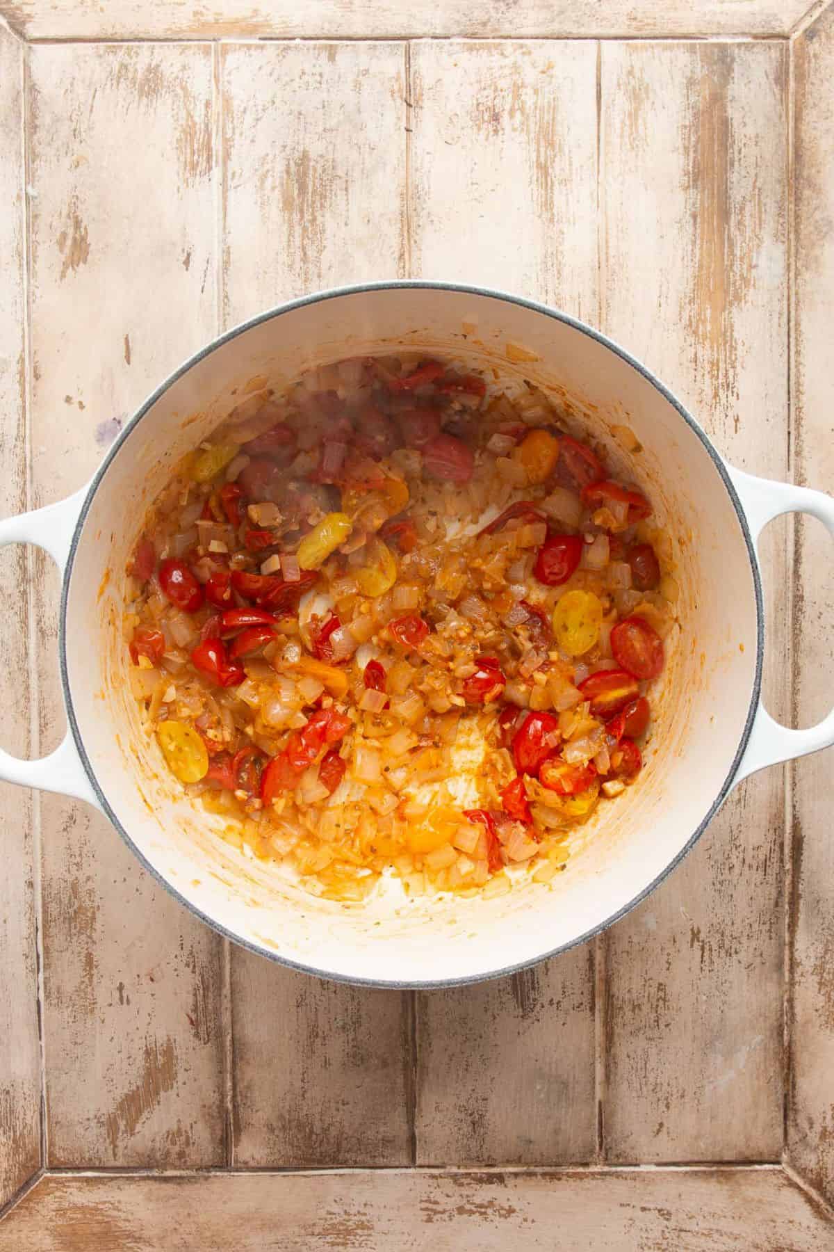 Halved red and yellow cherry tomatoes cooked down with diced onion and garlic in a white Dutch oven.