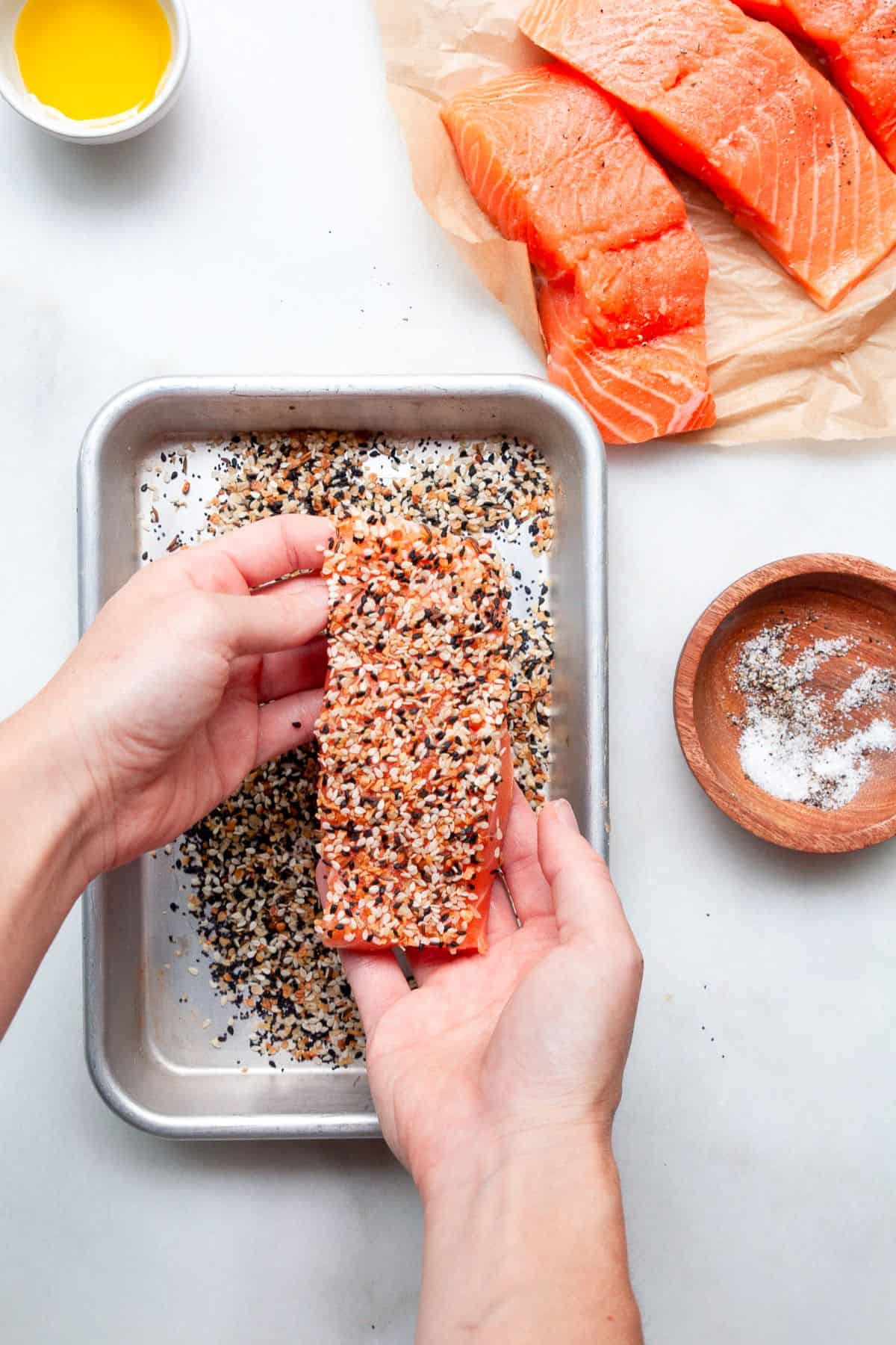 Hands pressing a salmon fillet into everything bagel seasoning on a sheet pan.