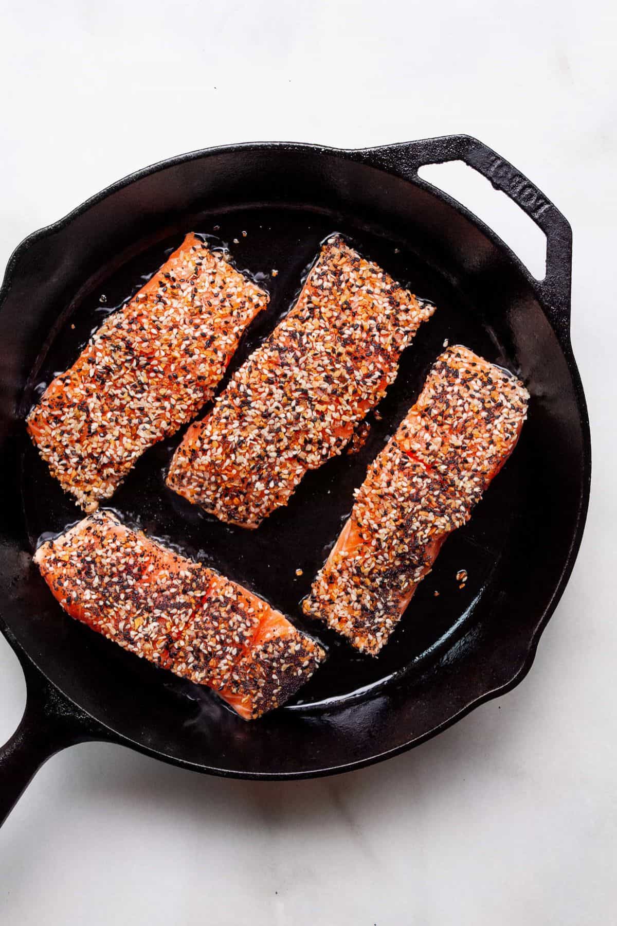 Four everything bagel crusted salmon fillets searing in a cast iron skillet.