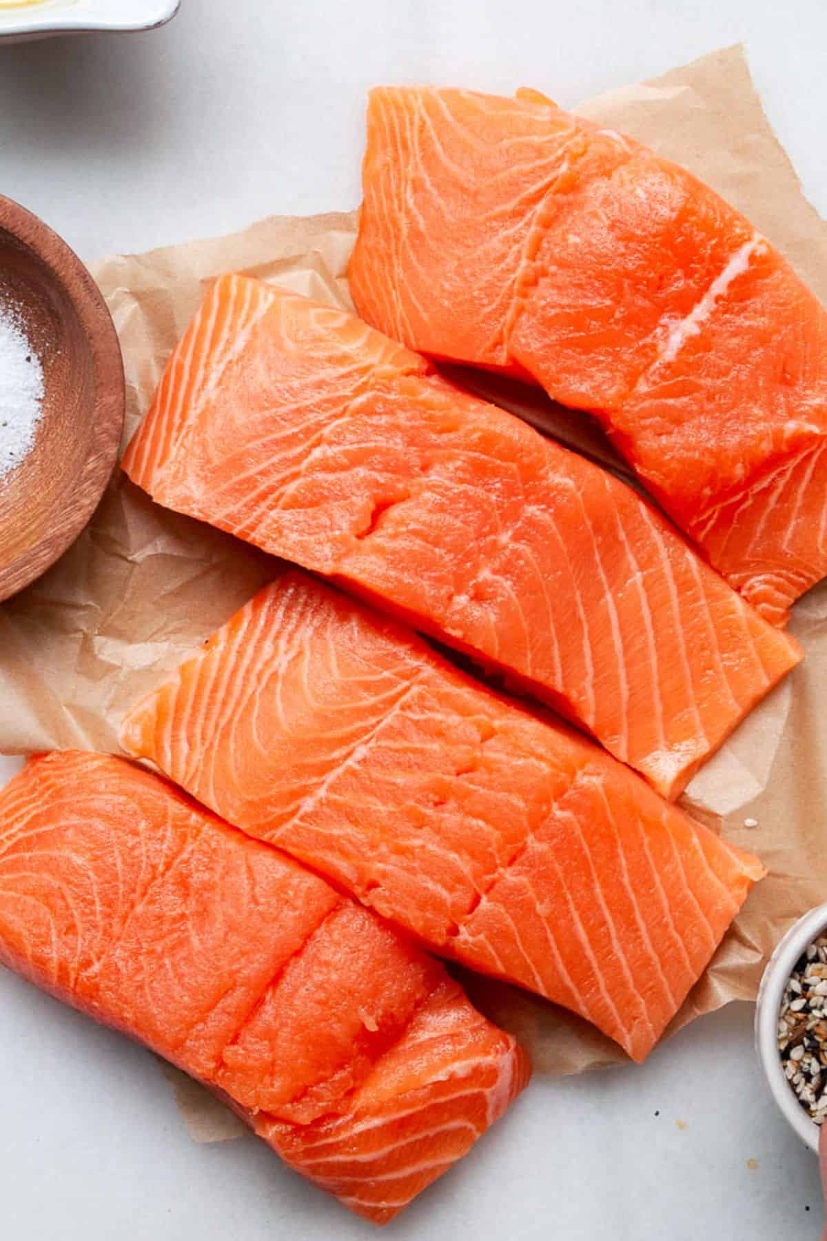 Four raw salmon fillets on parchment paper next to small bowls of salt and furikake seasoning.