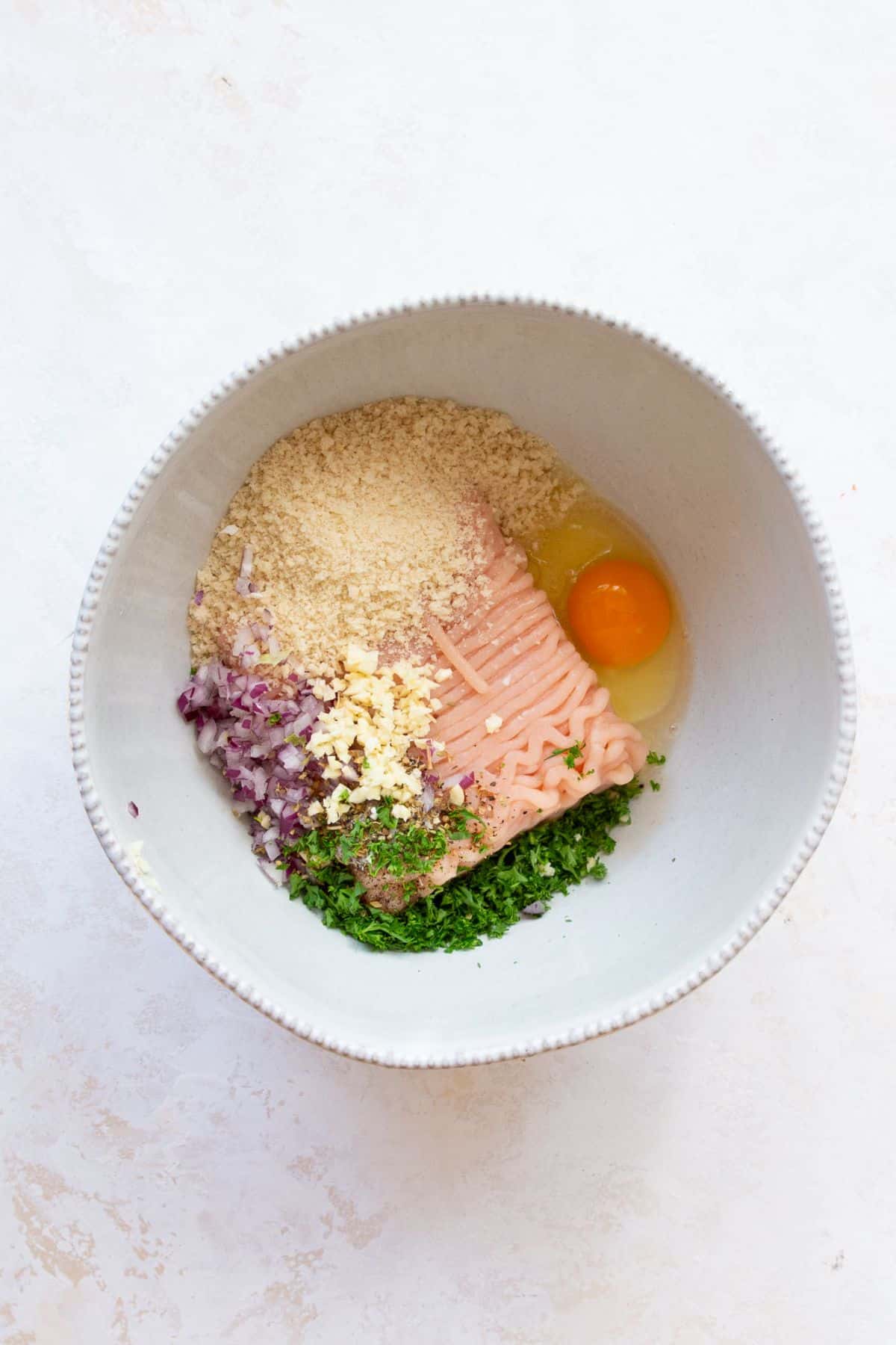 Ground chicken, panko, egg, diced red onion, minced garlic, and fresh parsley in a white mixing bowl before mixing.