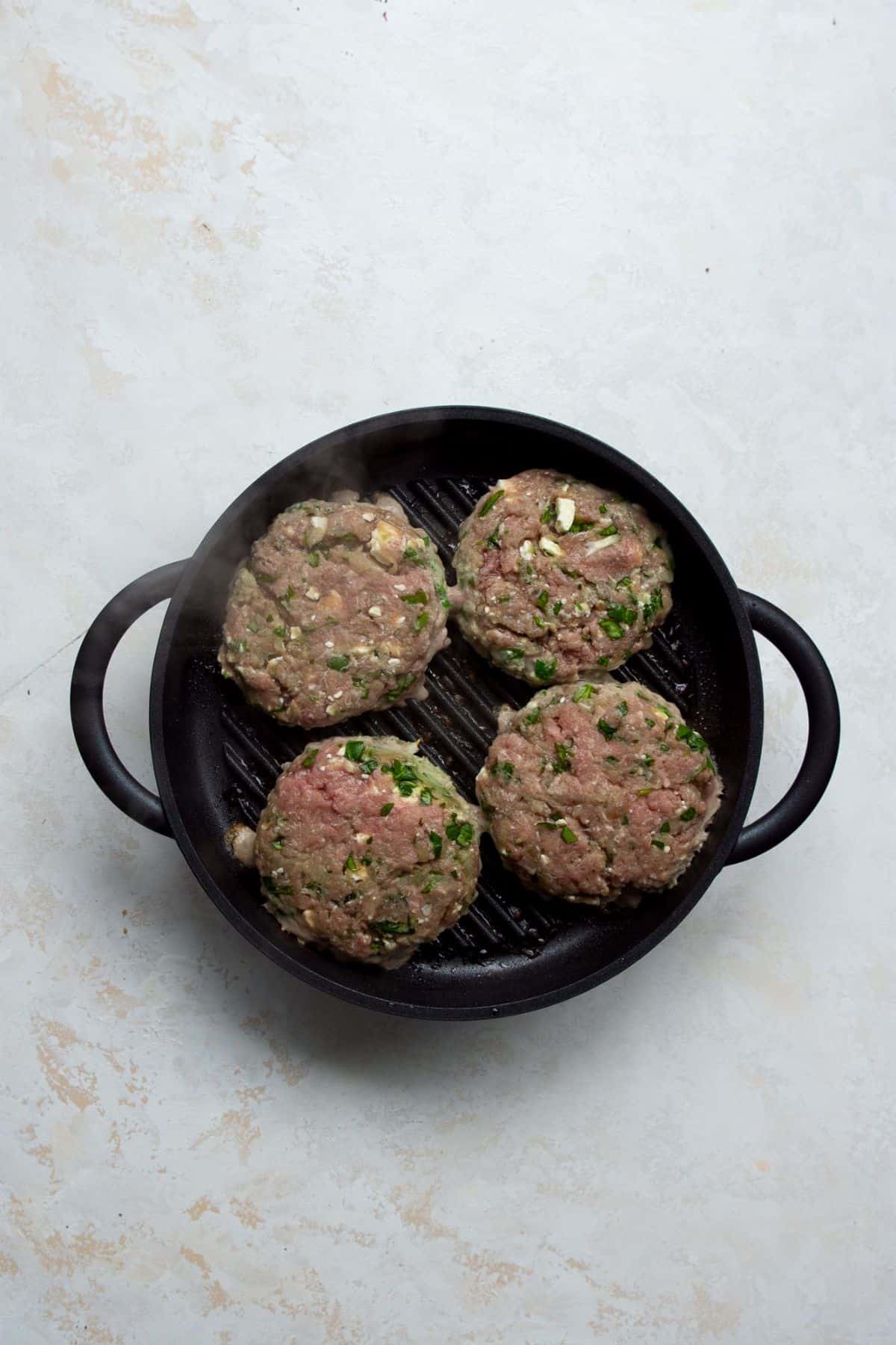 Four raw Greek turkey burger patties with visible feta and spinach on a hot grill pan.