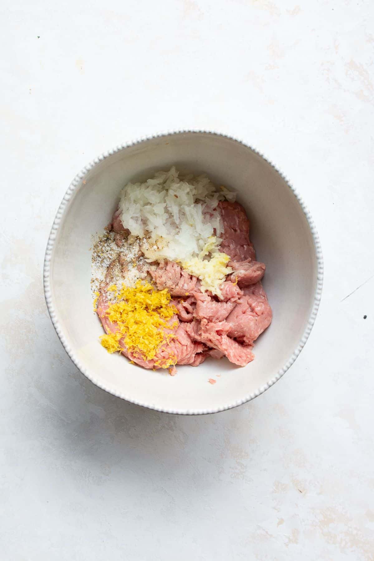 Ground turkey, shredded onion, lemon zest, and seasonings in a white bowl before mixing.