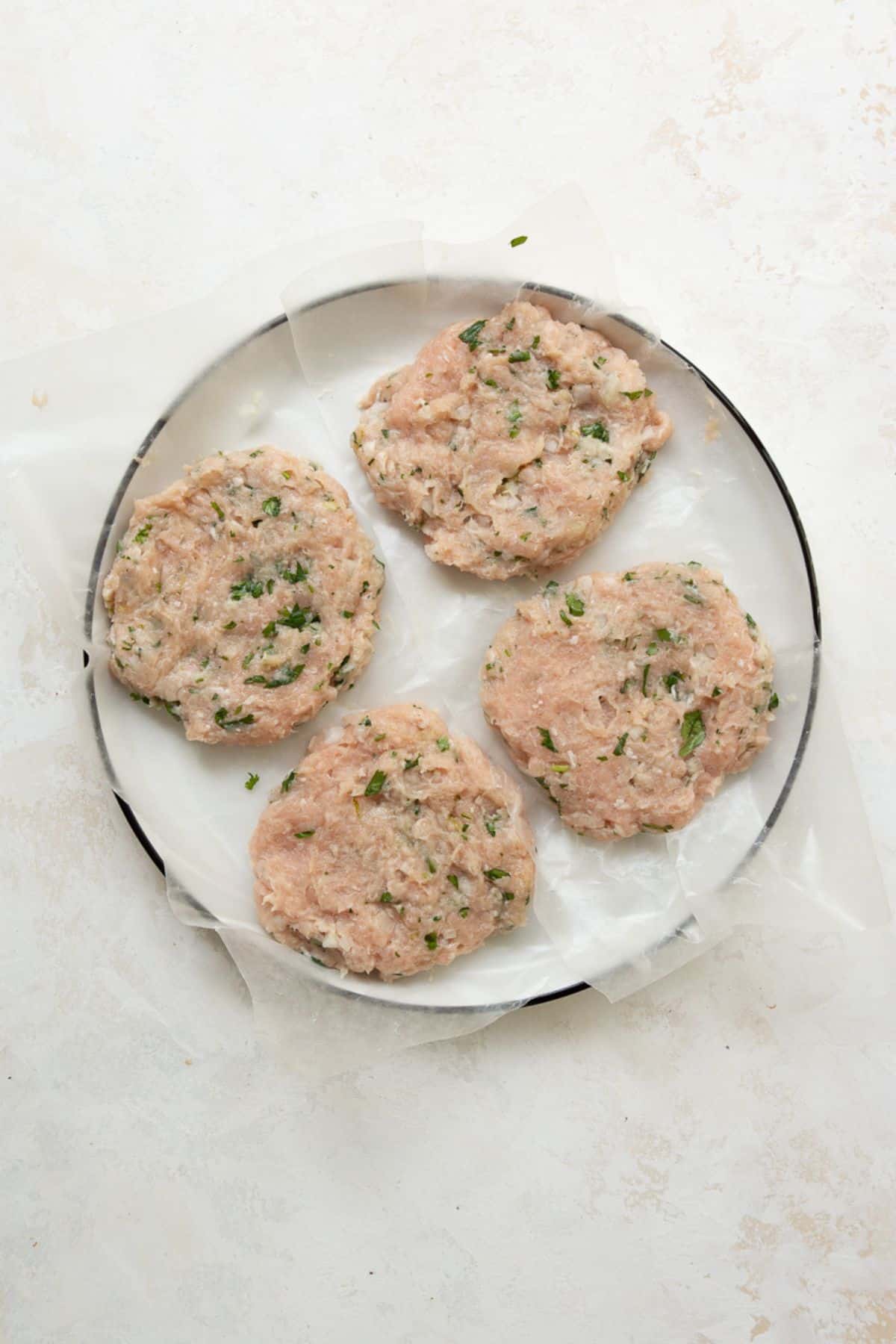 Four raw ground chicken burger patties on a parchment lined plate.