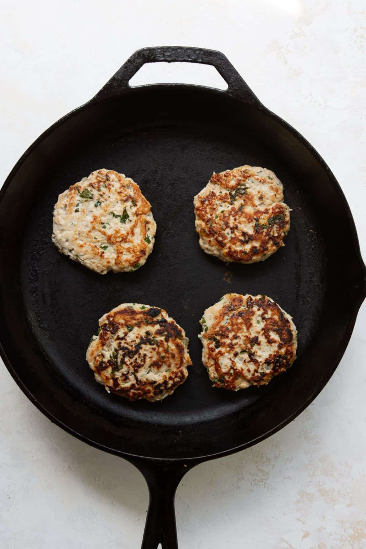 Four cilantro lime chicken burgers cooking in a cast iron skillet until golden brown.