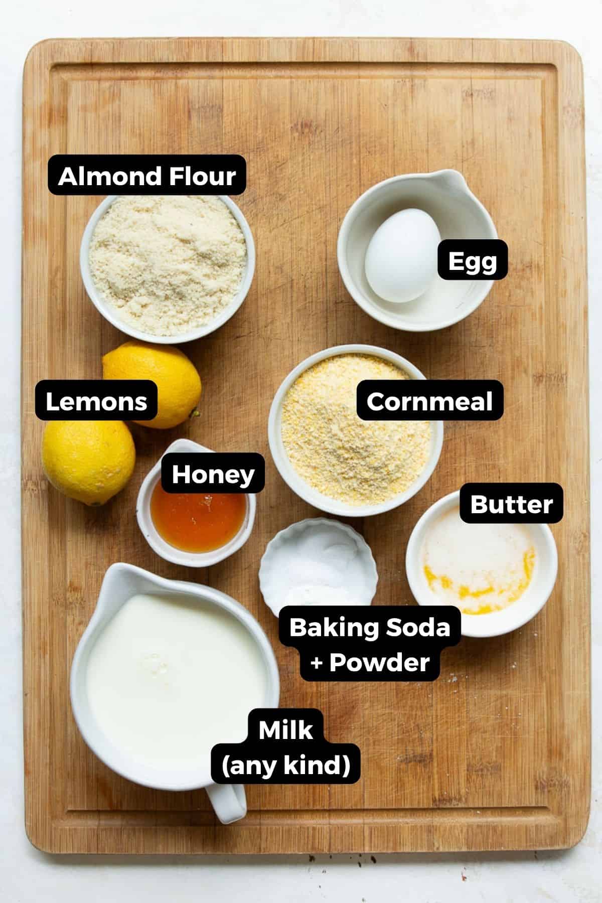 Ingredients for healthy almond flour cornbread measured into bowls on a wooden cutting board including almond flour, cornmeal, honey, melted butter, an egg, milk, and lemons.