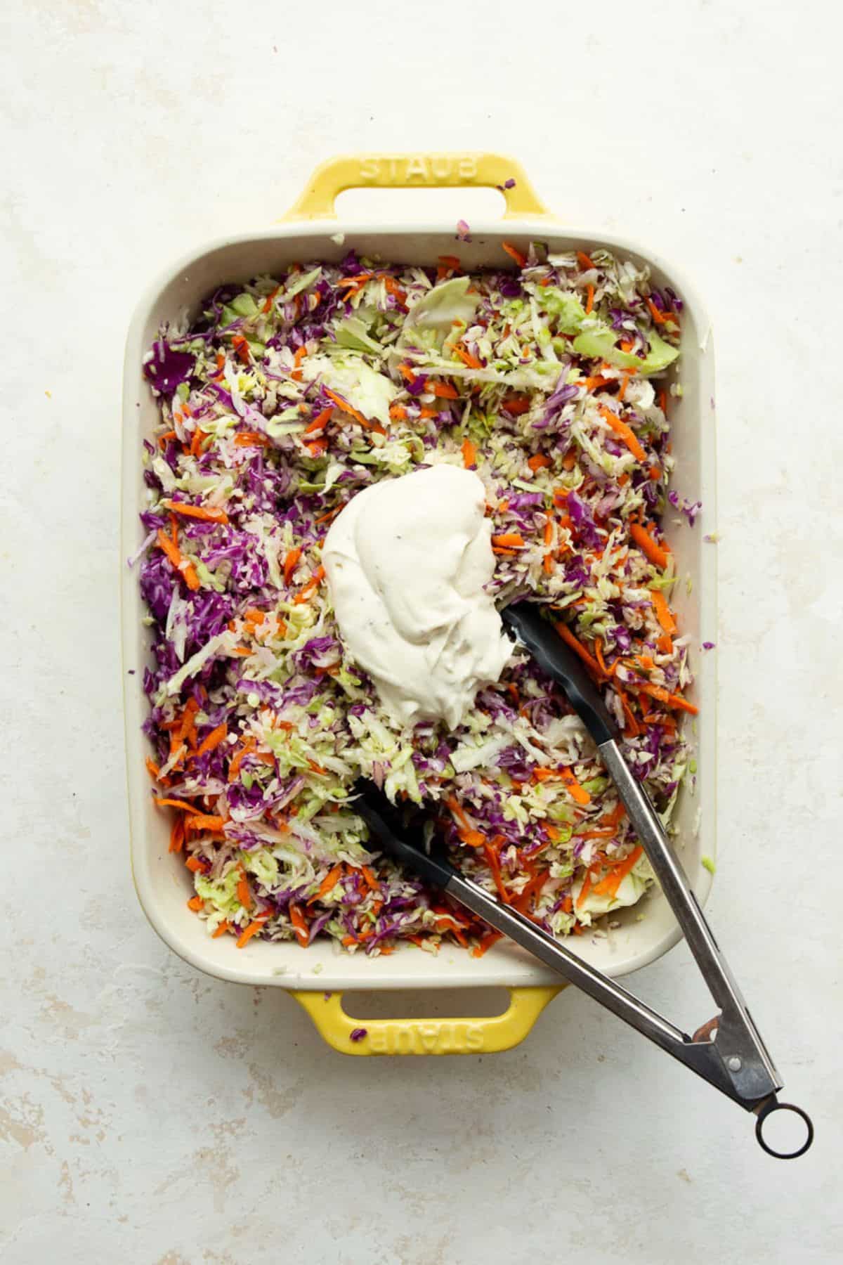 Creamy Greek yogurt dressing spooned onto the shredded cabbage and carrots with tongs resting in the dish.