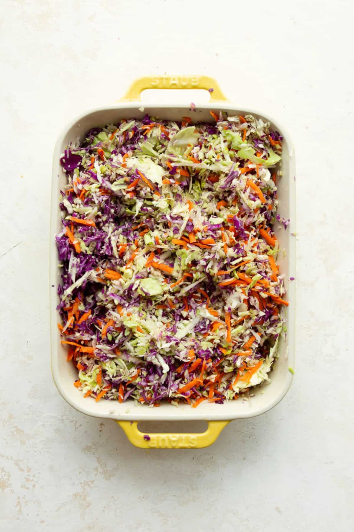 Shredded green cabbage, purple cabbage, carrots, and red onion tossed together in a white baking dish.