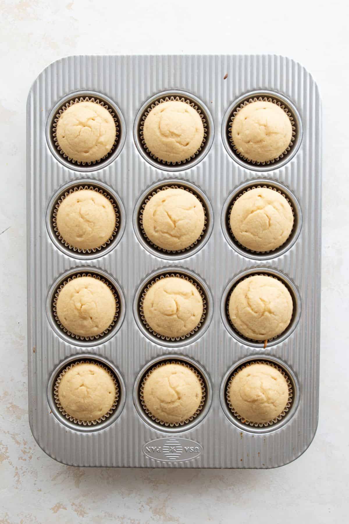 Twelve baked vanilla cupcakes cooling in the tin, domed and golden on top.