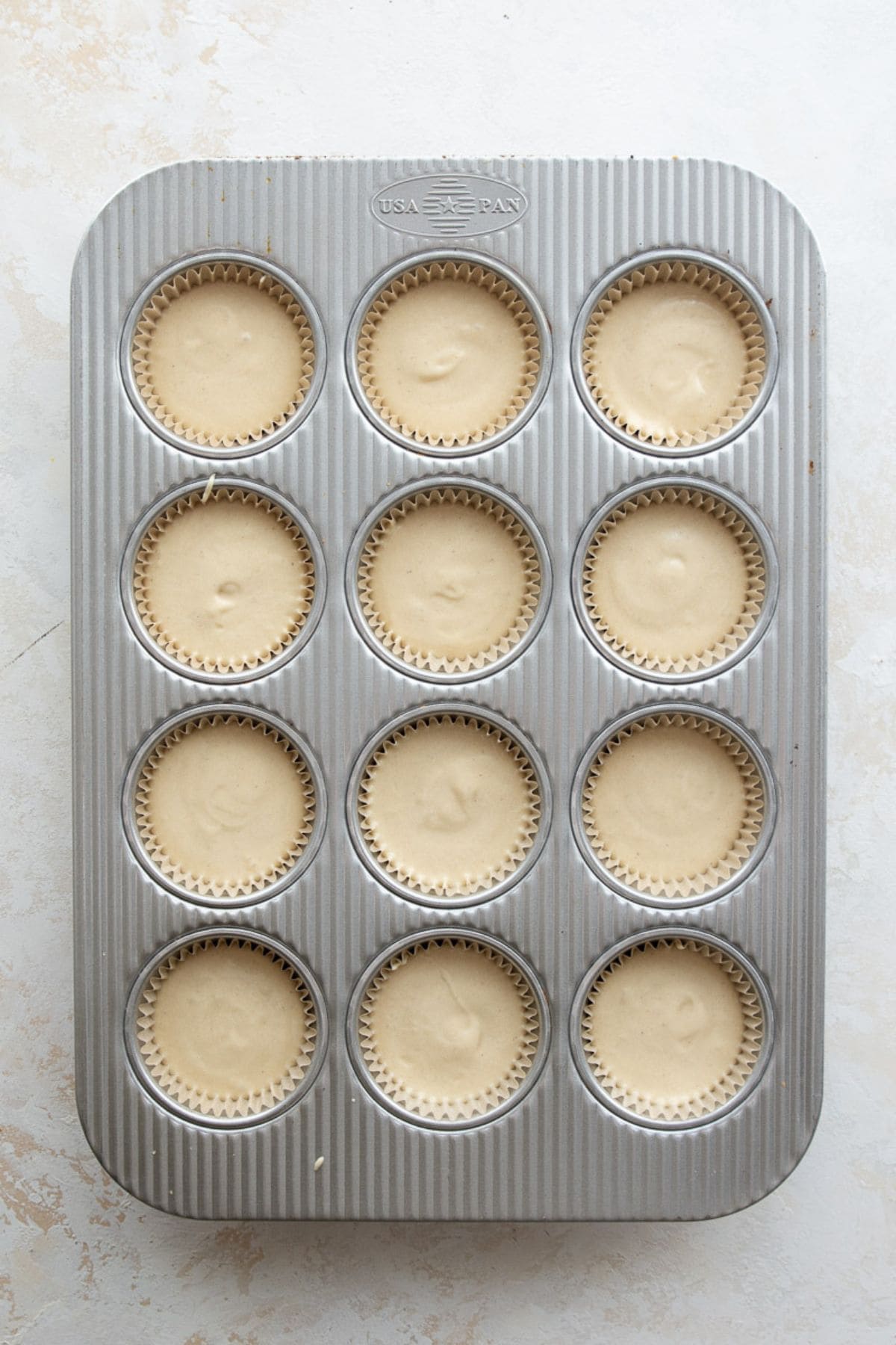 Twelve cupcake liners filled two-thirds full with vanilla batter in a metal cupcake tin.