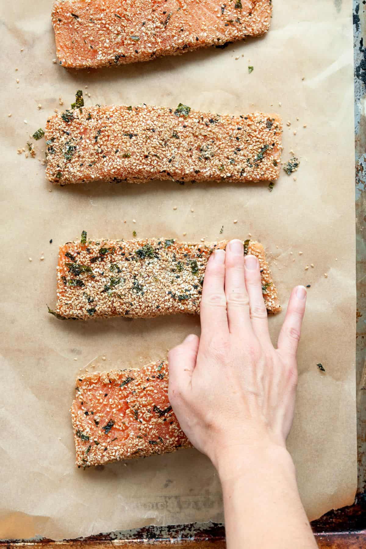 Hand pressing furikake seasoning onto four raw salmon fillets on a parchment lined baking sheet.